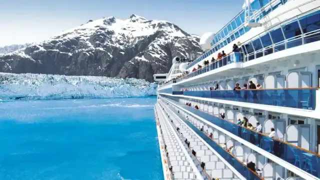 A cruise ship with passengers on deck sailing past a tidewater glacier and snowy Alaskan peaks