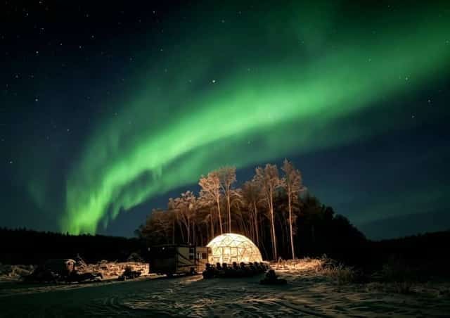 A glowing glass dome under a vivid green aurora borealis in a snowy Alaskan forest