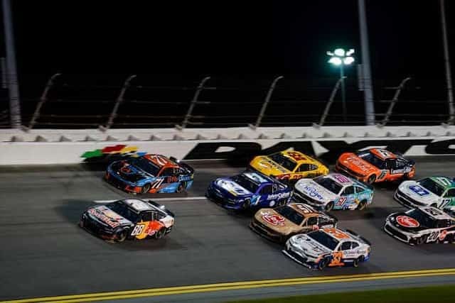 NASCAR stock cars racing door to door on a lit speedway at night, Daytona signage on the wall
