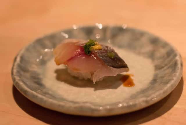 A single piece of aji mackerel nigiri garnished with herbs on a ceramic plate