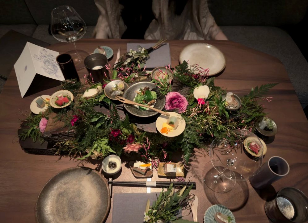 An elaborate kaiseki tasting spread arranged with fresh flowers and greens on a wood table